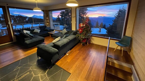 A modern living room with a large window offering a scenic view of mountains and a sunset. There are two black sofas, a coffee table, and a small Christmas tree. - Chalet Chamrousse 1750, 7 Pers, 3 ch, Terrasse, vue Magnifique sur le Vercors (Chamrousse)