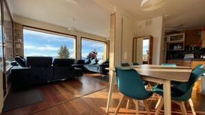 A modern living room with a sofa and chairs, a dining table with chairs, large windows with a view of the outdoors, and a kitchen area. - Chalet Chamrousse 1750, 7 Pers, 3 ch, Terrasse, vue Magnifique sur le Vercors (Chamrousse)