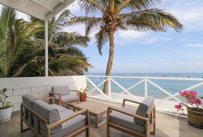 Un espace extérieur aménagé, abrité et doté de mobilier en bois et de plantes en pot, offrant une vue sur une plage où l’on peut apercevoir des bateaux et des palmiers.