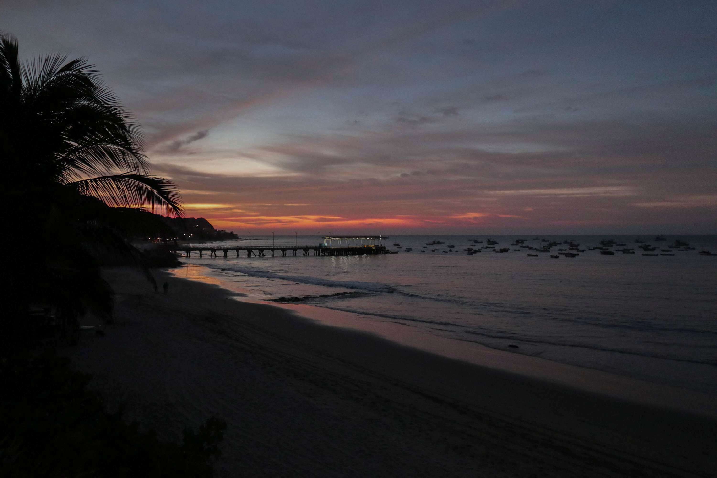 A beach with a pier extending into the water, palm trees, and a sunset.