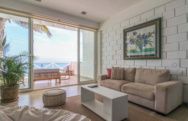 A modern living room with a beige sofa, a white coffee table, and a large sliding glass door leading to a balcony with a view of the ocean.