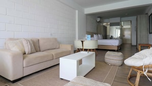 Una habitación de hotel moderna con un sofá color beige, una mesita de café blanca, un otoman pequeño y redondo, una cama con ropa de cama blanca y una mesita de noche de madera.