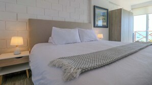 A modern hotel room with a large bed, white bedding, a woven throw blanket, a nightstand with a lamp, and a framed picture on the wall.