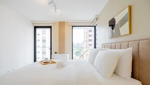 A hotel room with a large bed, a bedside table with a cup and a rolled towel, and a view of buildings through the window. - w* | Charming 1BR in San Francisco w/ View (Panamá)