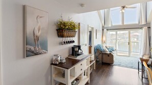 Een ruime kamer met een groot raam, een plafondventilator, een eethoek met een tafel en stoelen, en een wandplank met decoratieve objecten.