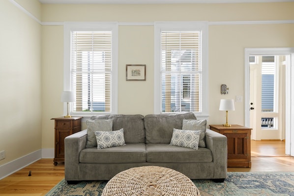 A living room with a gray sofa, patterned cushions, a woven ottoman, and a blue patterned rug.