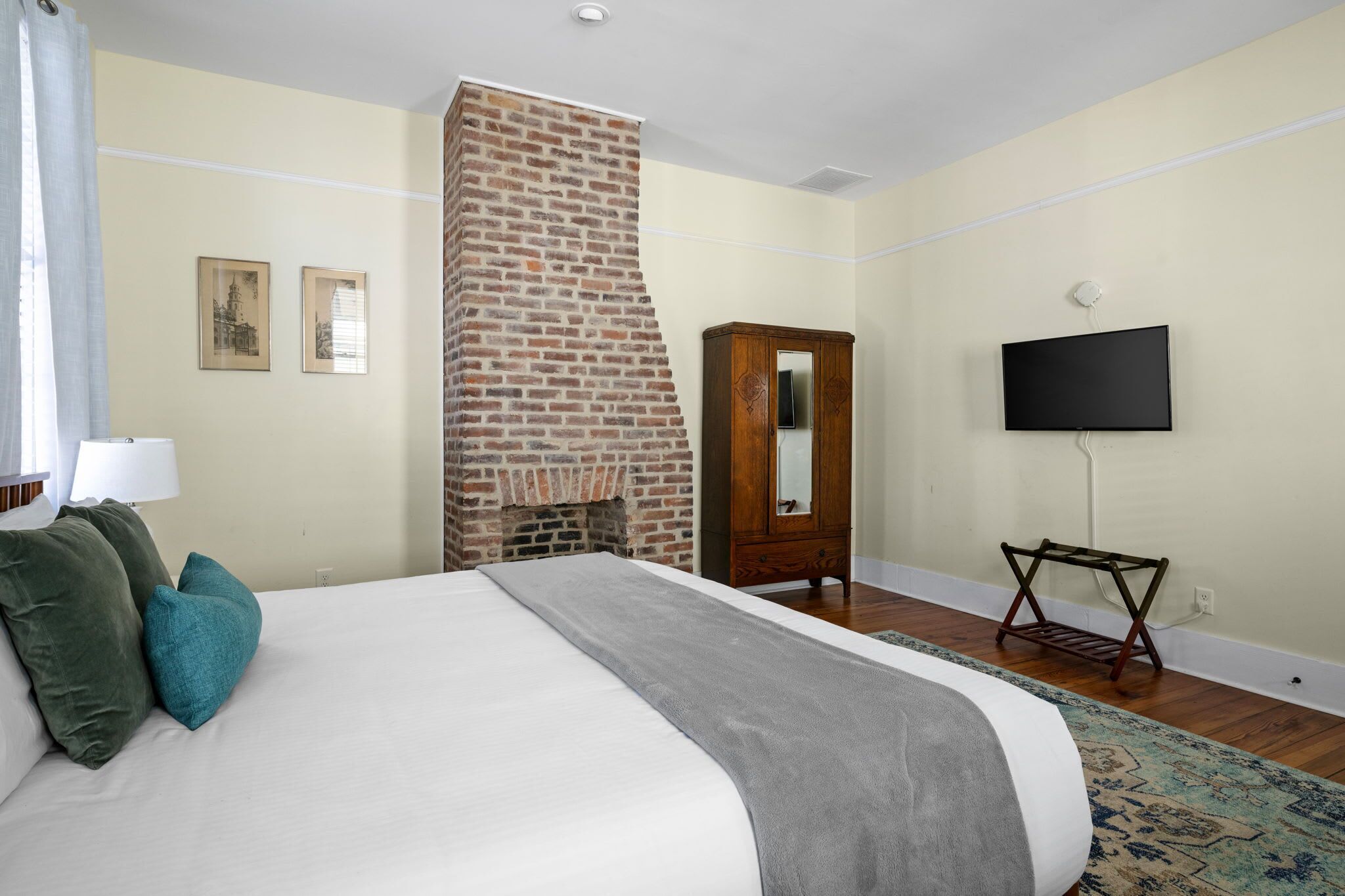 A bedroom with a brick fireplace, a bed with pillows, a wooden wardrobe, a television, and a patterned rug. - Edisto & Cooper House | Sleeps 14 | Full House (Charleston)