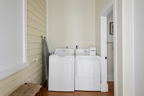 Une buanderie équipée de deux machines à laver à chargement frontal blanches et d’un sèche-linge, d’un banc en bois et d’une porte donnant sur une autre pièce.