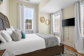 A bedroom with a bed, white curtains, a flat-screen TV, and two framed pictures on the wall.