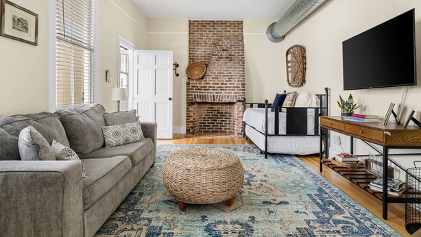 A living room with a grey sofa, a wicker ottoman, a coffee table, a TV, and a brick fireplace. - Edisto & Cooper House | Sleeps 14 | Full House (Charleston)