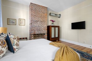 A hotel room with a brick fireplace, a flat-screen TV, a bed with floral pillows, and a wooden cabinet. - Edisto House | Great for Group Stays | Nightlife (Charleston)