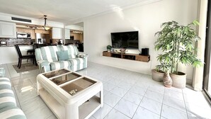 A modern living room with a striped sofa, a glass coffee table, a TV on a wooden shelf, and a large potted plant.