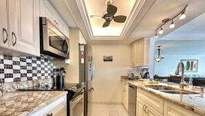 A modern kitchen with a microwave, oven, and stainless steel appliances. There is a ceiling fan and recessed lighting. The kitchen has a tiled backsplash and a granite countertop.
