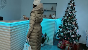 A person in a long beige coat standing in a room with a Christmas tree and presents.