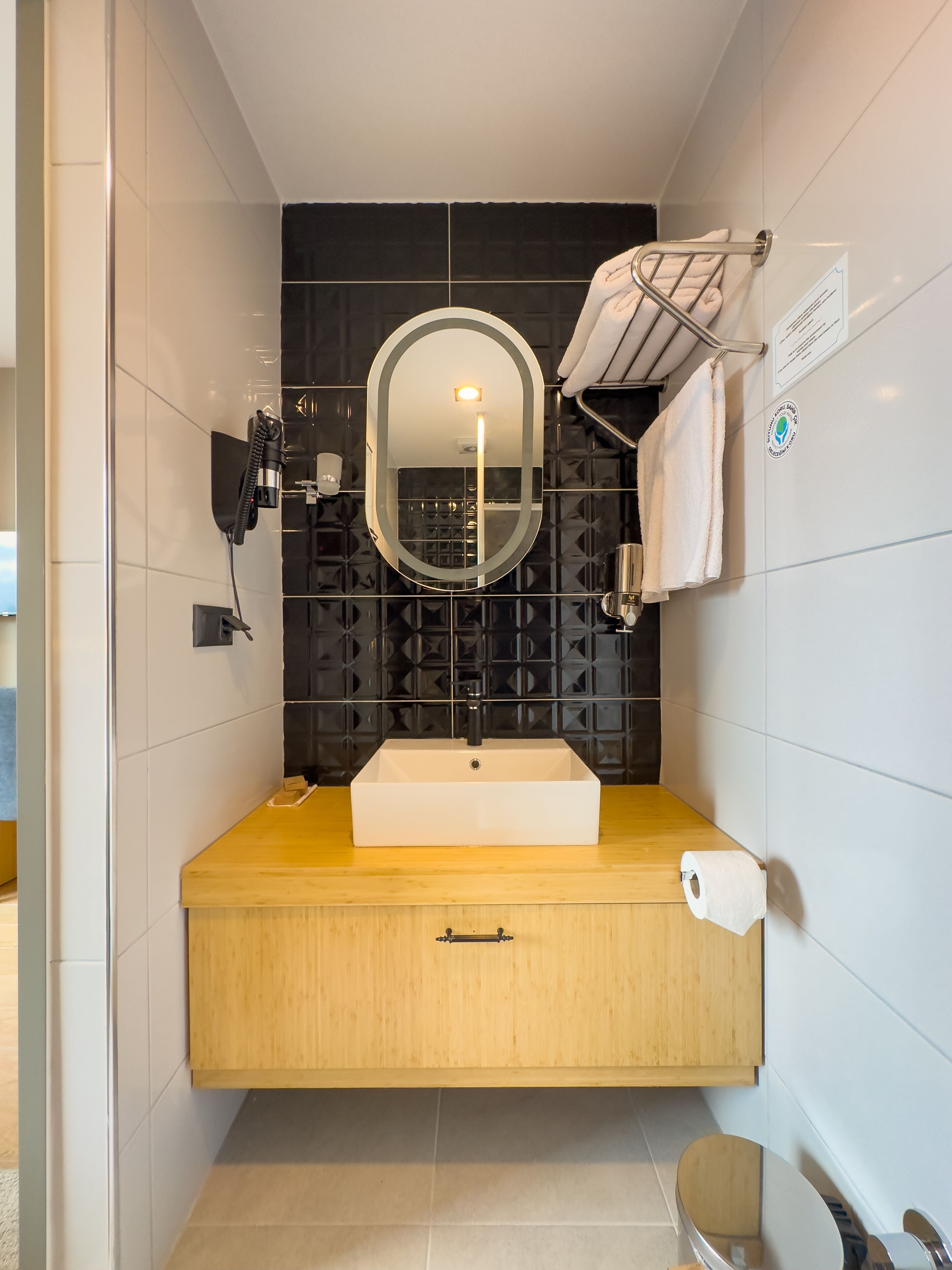 A modern bathroom with a rectangular sink, a mirror with integrated lighting, a towel rack, and a toilet paper holder.
