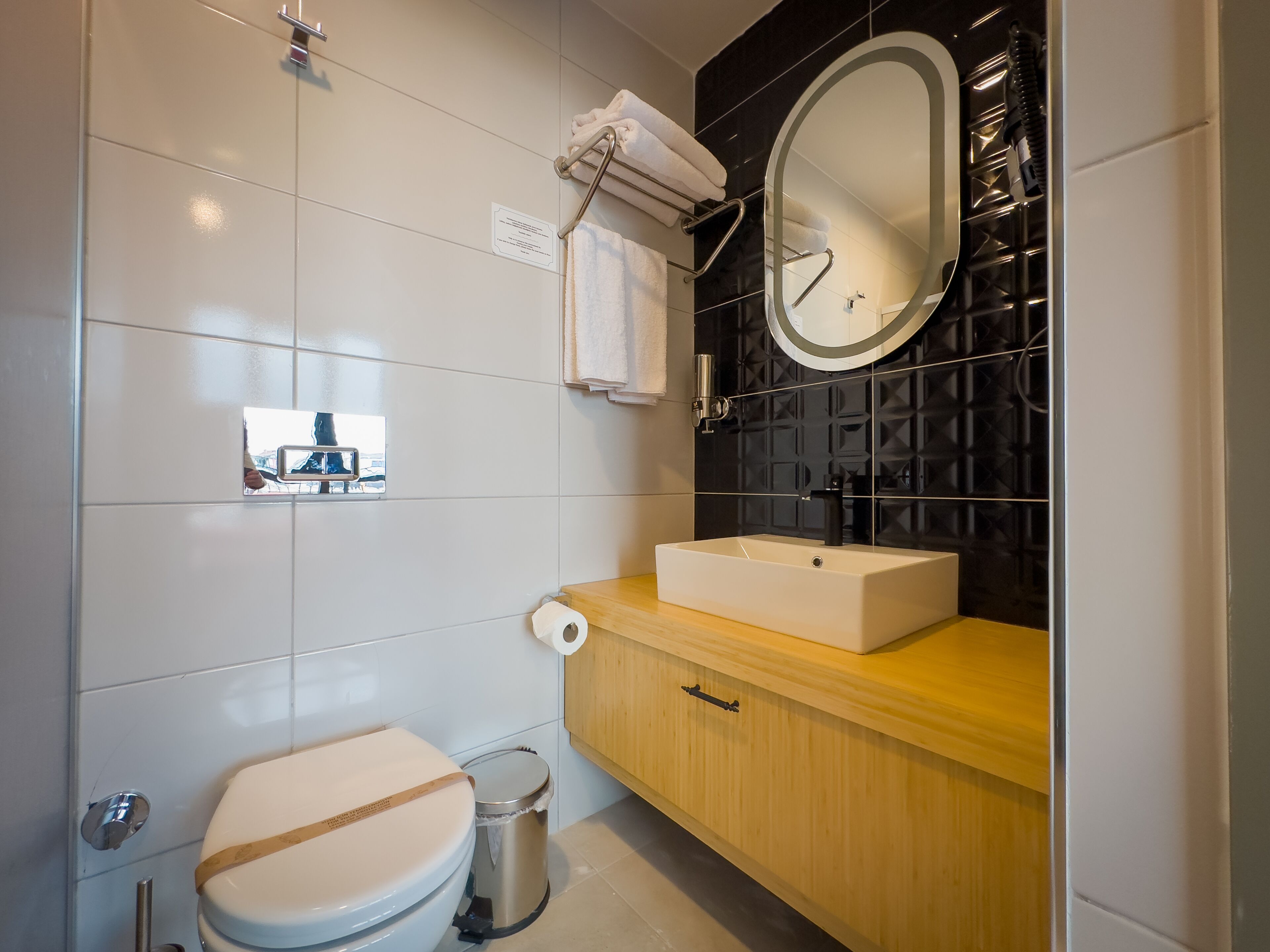 A modern bathroom with a white toilet, a wooden vanity with a sink, a mirror, and a black tiled wall.