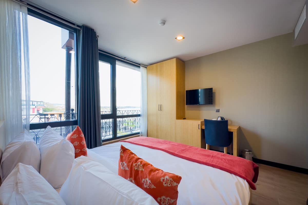 A hotel room with a large bed, a desk, a chair, a TV, and a balcony with a view.