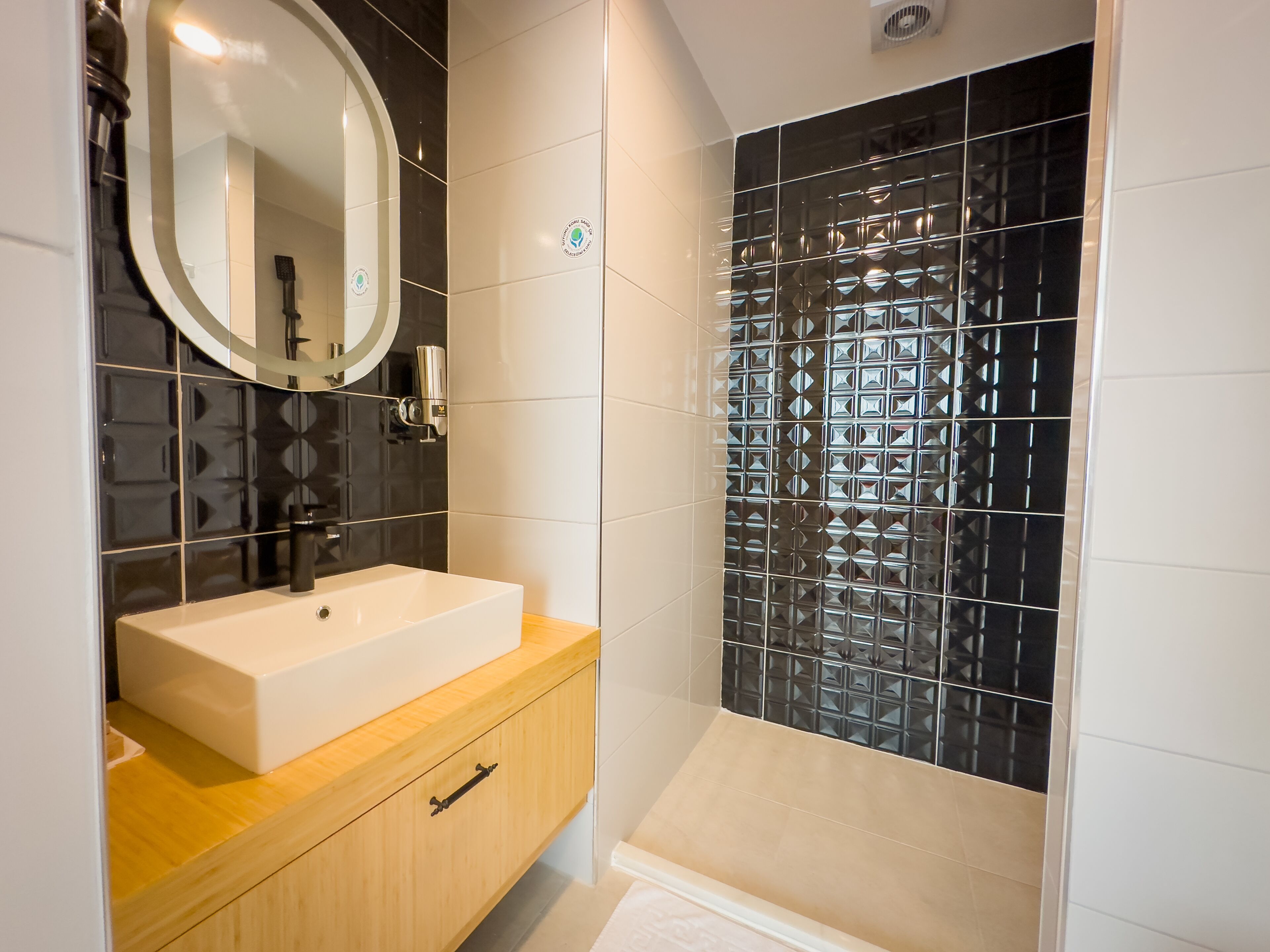Un baño con lavamanos blanco, tocador de madera y ducha con azulejos negros estampados.