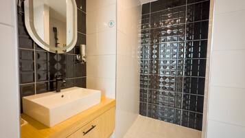 Un baño con lavamanos blanco, tocador de madera y ducha con azulejos negros estampados.