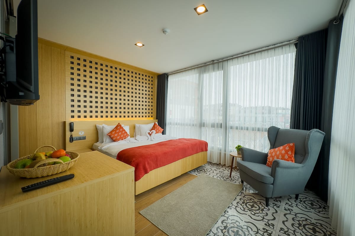 A hotel room with a bed, a desk with a basket of fruit, a chair, and a large window with curtains.