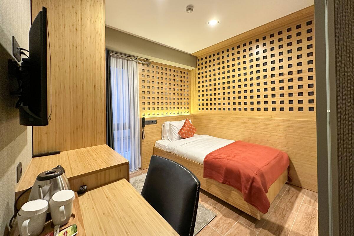A compact hotel room with a bed, a desk with a chair, a TV mounted on the wall, and a wooden partition with a grid pattern.