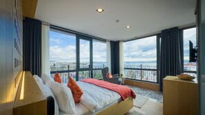 A modern bedroom with a large bed, a chair, and a view of a cityscape through large windows. - Mihman Tuzla (Tuzla)