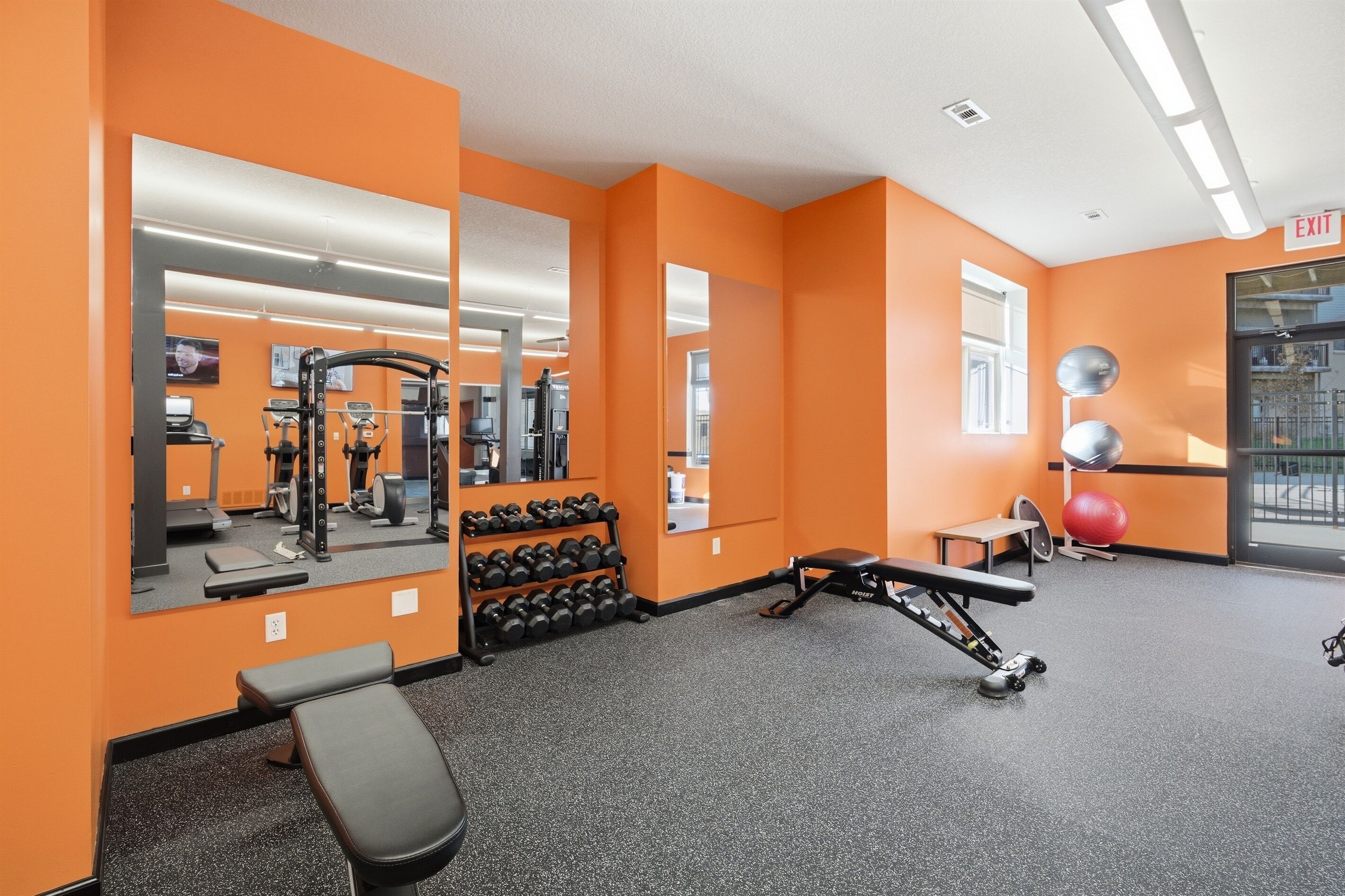 Een sportschool met oranje wanden, trainingsapparatuur en een grote spiegel.