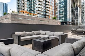 A rooftop terrace with a modern outdoor seating area, including a sectional sofa and a fire pit table, overlooking a cityscape with high-rise buildings.