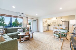 Un amplio salón con sofá, una mesa de centro y una mesa de comedor. Se observa una cocina al fondo con armarios blancos y un refrigerador.