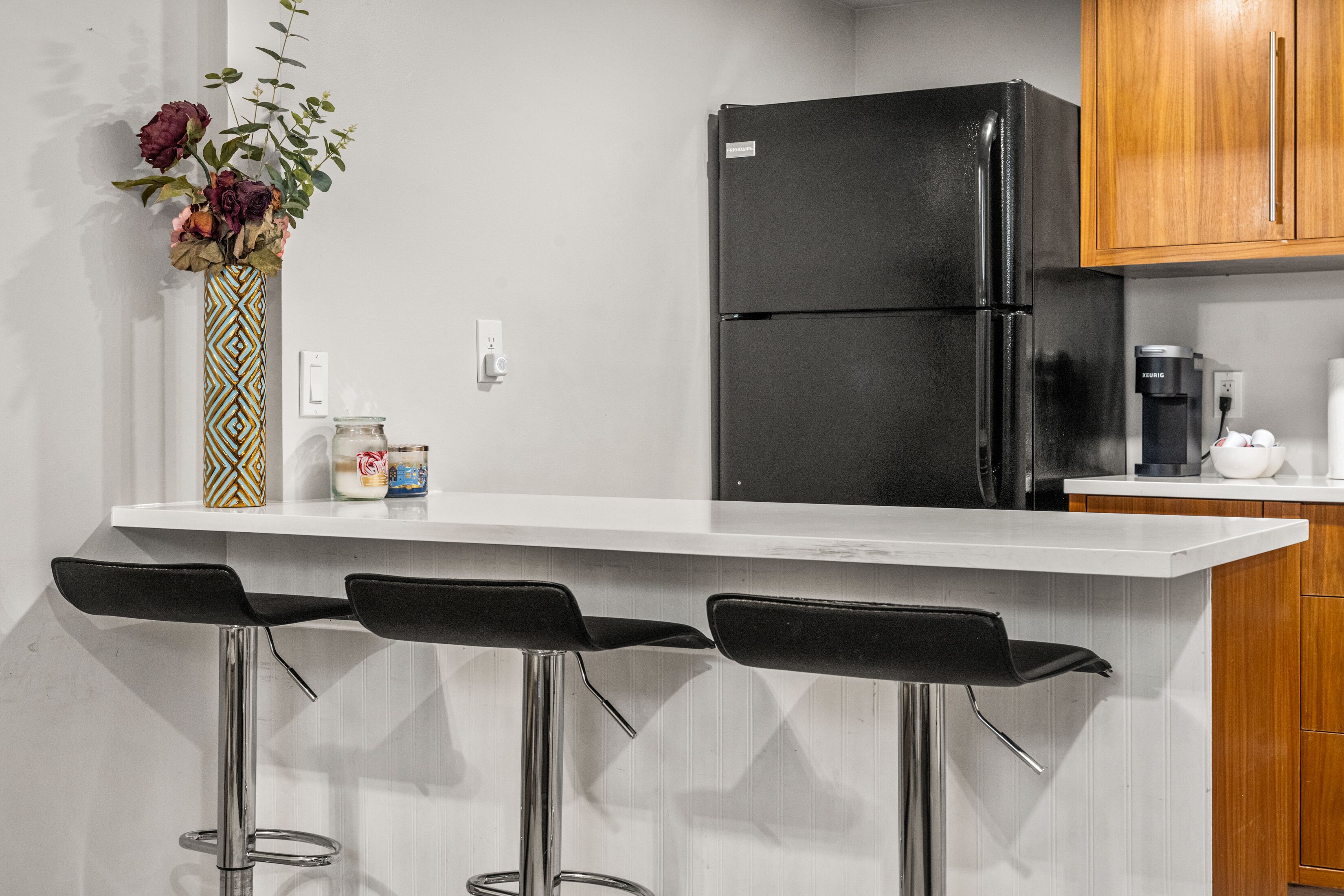Una cocina con un refrigerador negro, armarios de madera, una encimera blanca y tres taburetes negros.