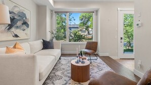 Uma sala de estar moderna com um sofá branco, uma poltrona marrom, uma mesa de centro redonda de madeira e uma grande janela com vista para vegetação e casas.