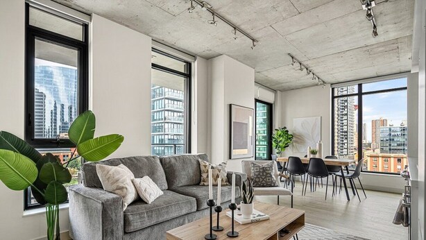 Uma sala de estar moderna com um sofá cinzento, uma mesa de centro de madeira e uma área de jantar com mesa e cadeiras. Grandes janelas oferecem vistas da paisagem urbana.