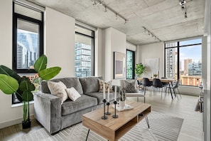 A modern living room with a grey sofa, a wooden coffee table, and a dining area with a table and chairs. Large windows provide views of the cityscape.
