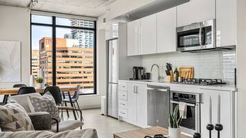 A modern kitchen with white cabinets, a stainless steel microwave, and a dishwasher. There is a dining area with a wooden table and chairs, and a large window offering a view of a building outside.