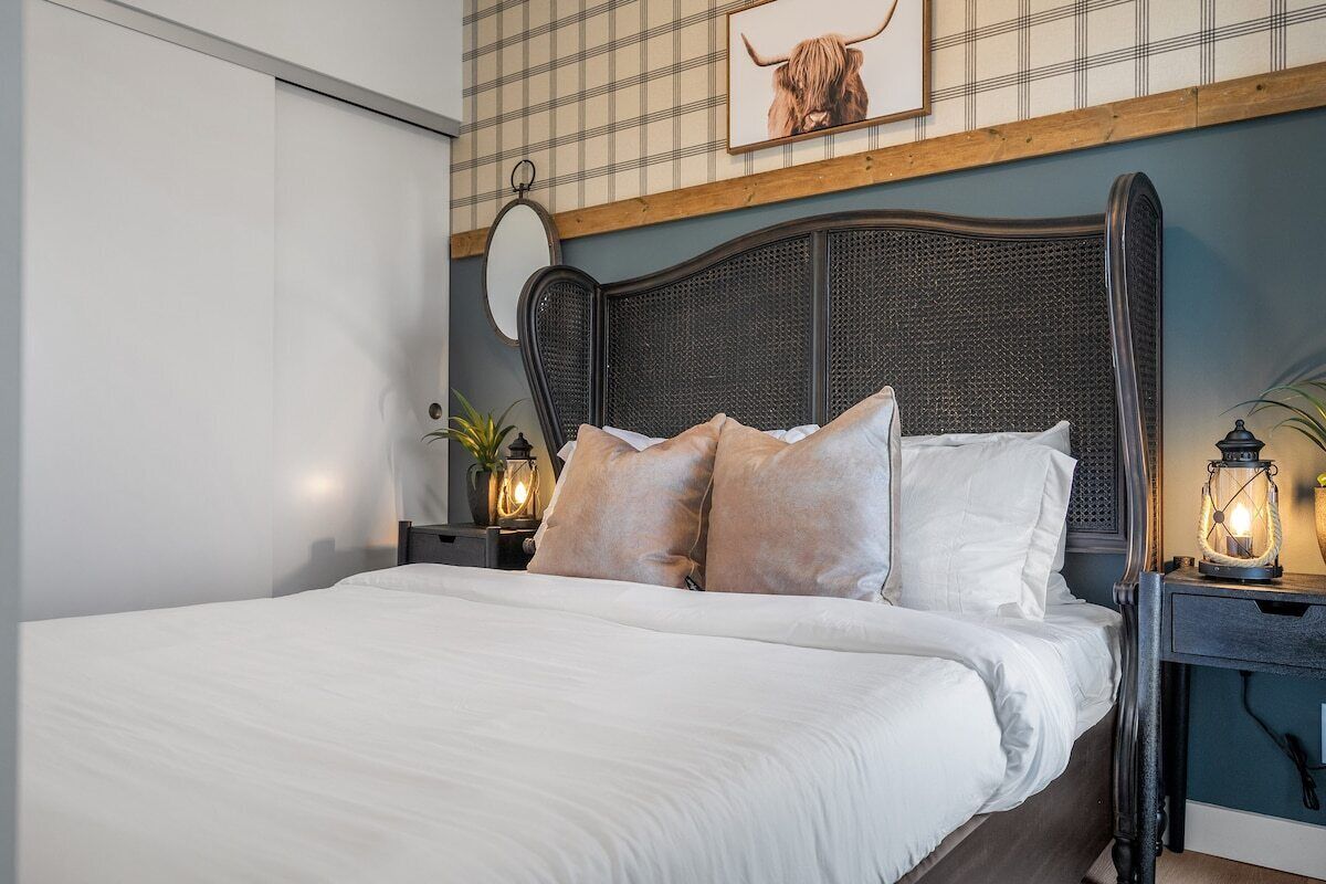 A neatly made bed with white linens and two pillows, a wooden headboard, a wall-mounted mirror, and a framed picture above the bed.