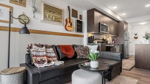 A modern living room with a black leather sofa, a round coffee table, and a small side table. A kitchen is visible in the background with wooden cabinets and modern appliances. - Howdy! 1BR Western Theme w Rooftop Patio & AC (Calgary)