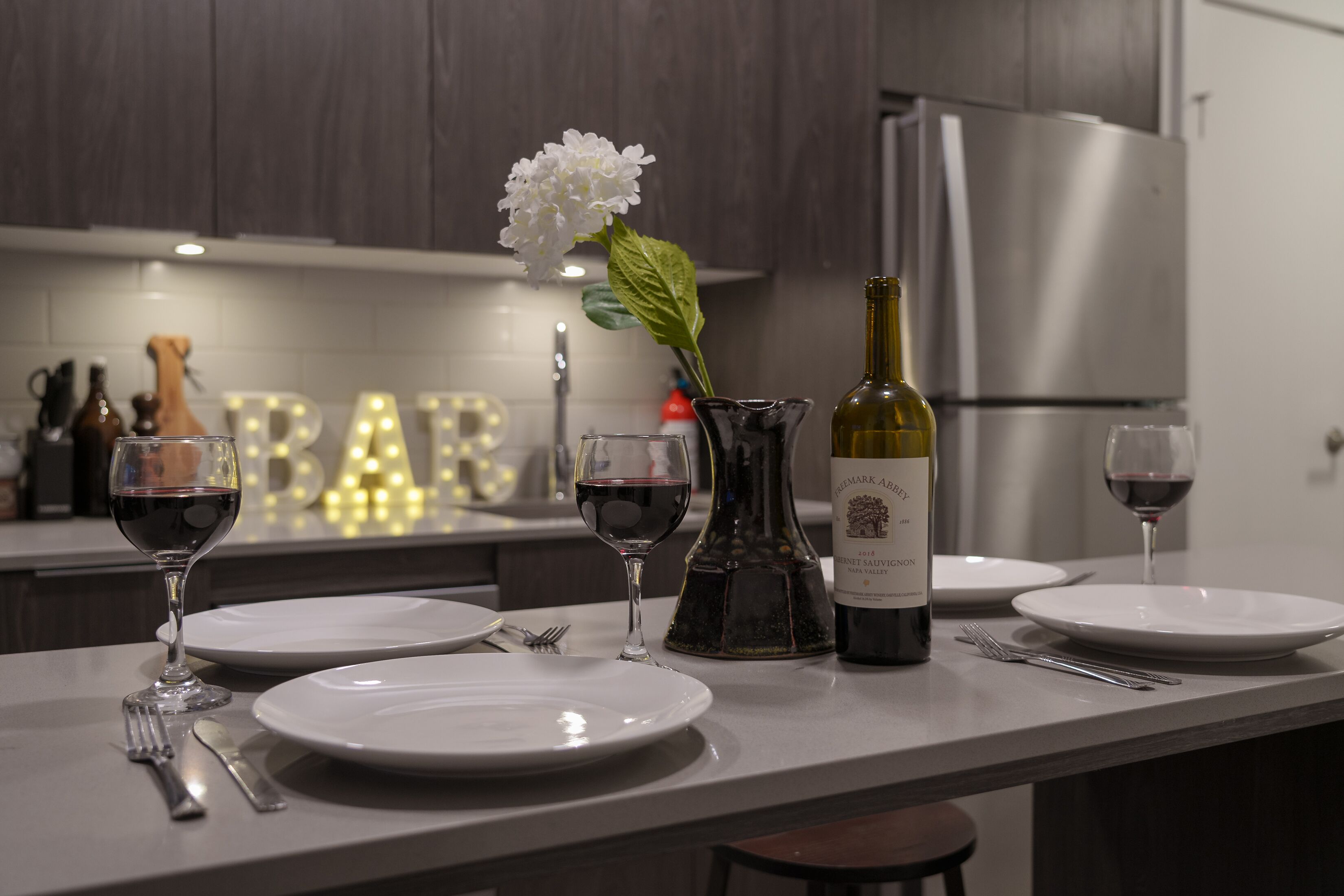 Una cocina moderna con una mesa de comedor para dos personas, con copas de vino, una botella de vino y un florero con una flor blanca.