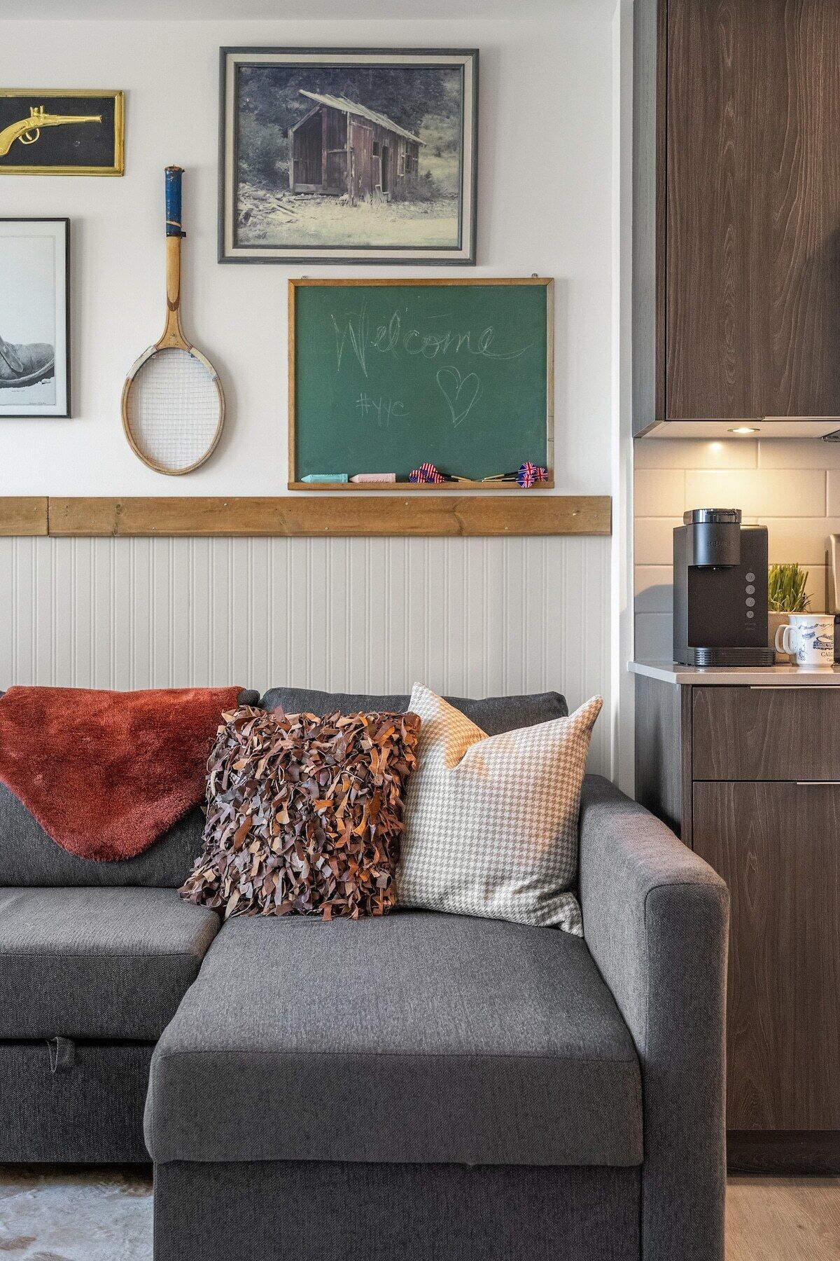 A cozy living area with a grey sofa, a red throw blanket, and decorative pillows. A chalkboard with "Welcome" and a heart is mounted on the wall above the sofa.