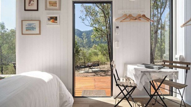 Una camera da letto con un letto, un tavolo con tovaglia e una sedia. Alle pareti ci sono quadri incorniciati, e si gode una vista sull'esterno attraverso una porta.