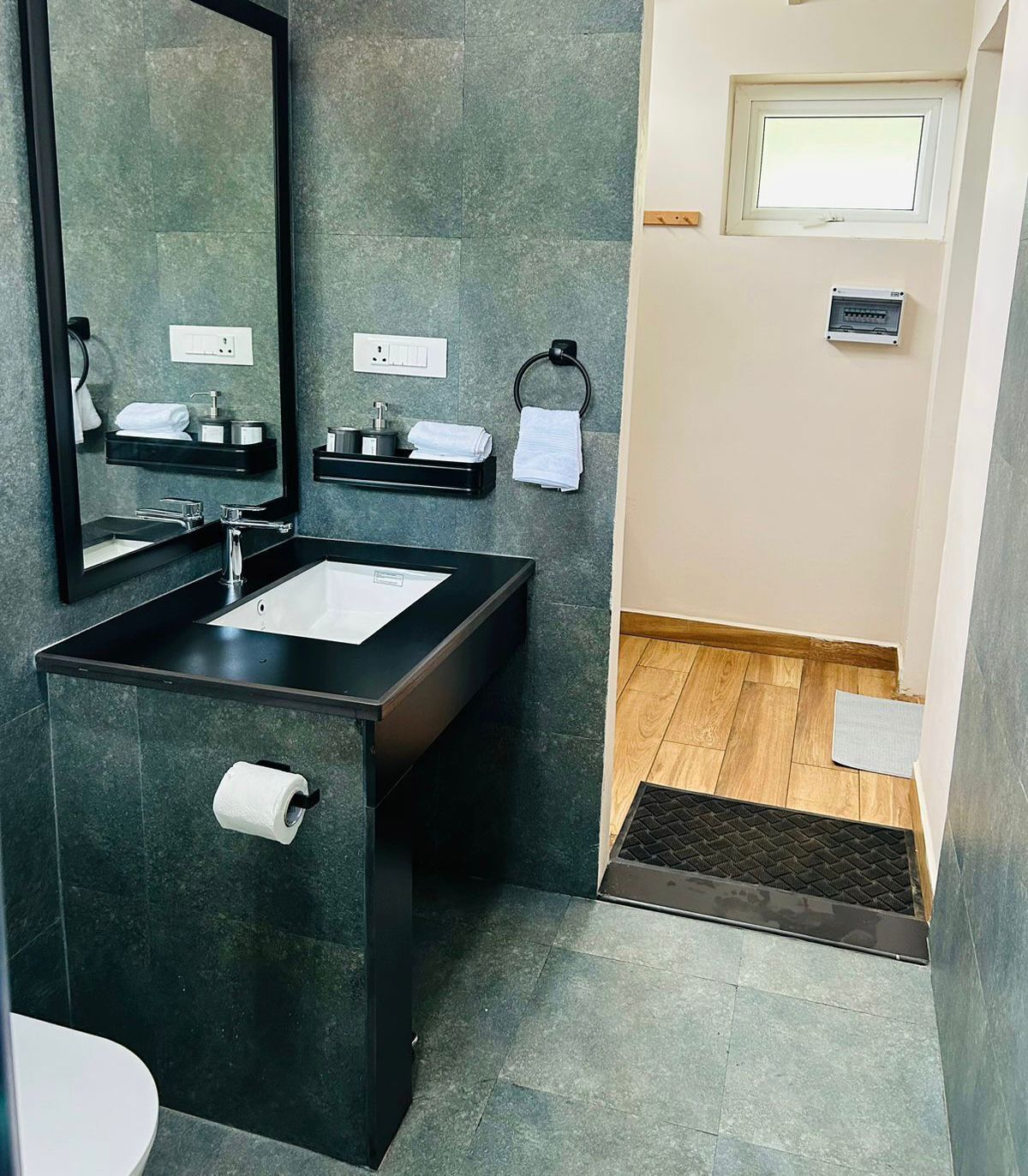 A modern bathroom with a black sink, mirror, and towel rack. There is a window above the sink and a door with a mat in front.