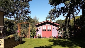 Une petite maison de plain-pied, avec un toit rouge et une porte violette, entourée d’un jardin bien entretenu où poussent différentes espèces de plantes et délimitée par une clôture en bois.