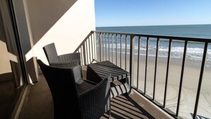 A balcony with two wicker chairs and a small table, overlooking a beach with waves and a clear sky.