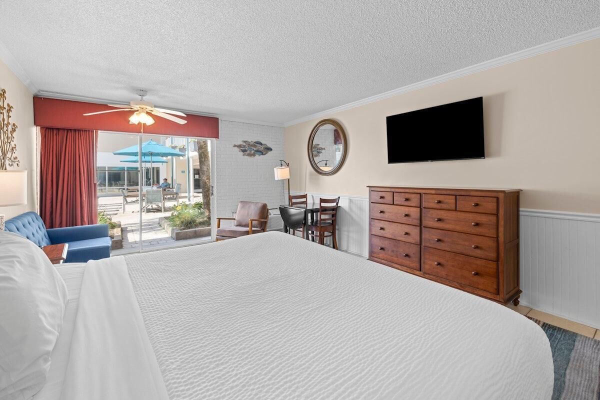 Une chambre d’hôtel dotée d’un grand lit, d’une commode en bois, d’un bureau avec des chaises, d’une télévision et d’un balcon avec des sièges d’extérieur.