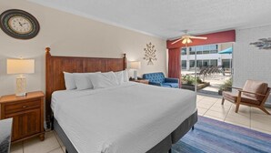 A hotel room with a large bed, a wooden headboard, a nightstand with a lamp, a blue armchair, a ceiling fan, and a view of the outside. - Hidden Treasures, 1st Floor, Pool View King Room (Pawleys Island)