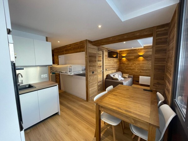 Cocina compacta con pileta, estufa y alacenas blancas. Hay un comedor con una mesa y sillas de madera.