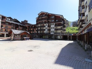 Una piazza con lastricato di ciottoli, circondata da edifici a più piani con balconi, una struttura in legno con tetto spiovente e un cartello di una caffetteria.