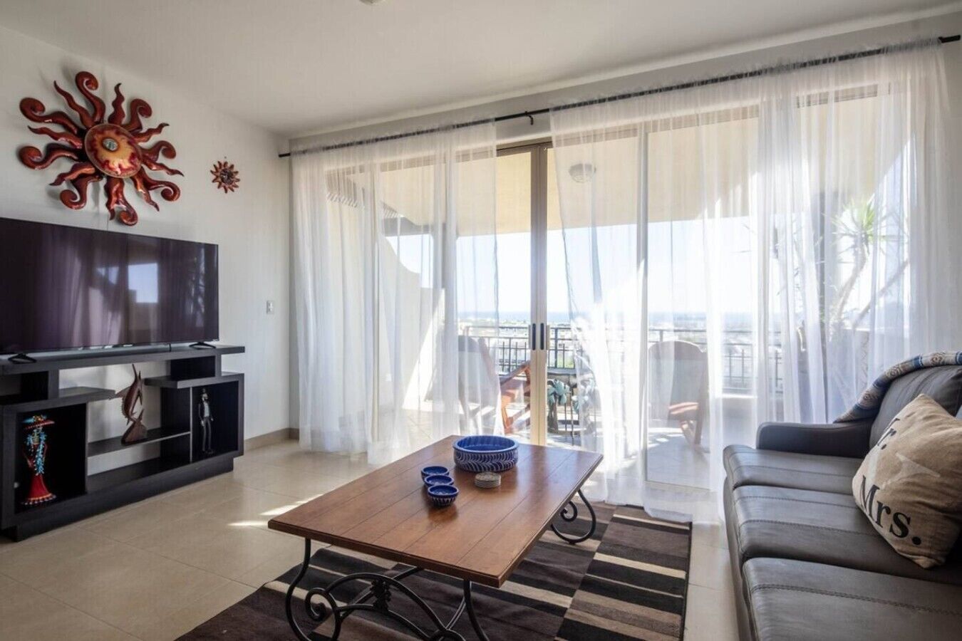 Ein Wohnzimmer mit Flachbildfernseher, einem Couchtisch mit dekorativen Schalen, einer Couch mit einem Kissen mit der Aufschrift 'Mrs.' und einem Balkon mit transparenten Vorhängen.