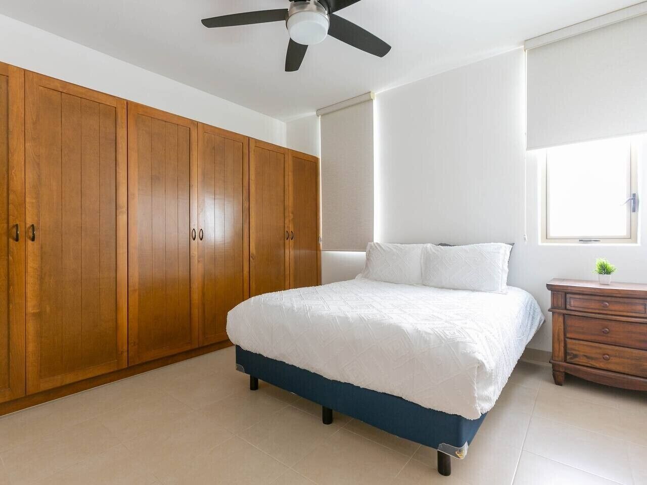 Een slaapkamer met een bed, een houten garderobe, plafondventilator en een raam met zonwering.