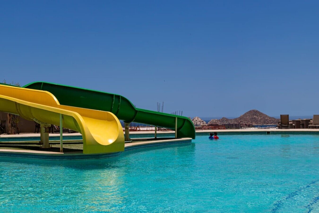 A swimming pool with two slides, clear blue water, and a distant view of a mountainous landscape.
