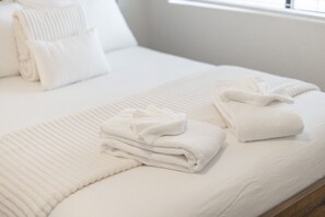 Un lit bien fait, avec du linge de lit blanc et des oreillers, une serviette pliée posée sur le lit, et une fenêtre en arrière-plan.